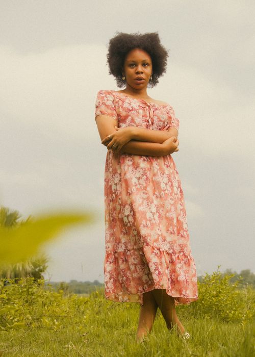 Free Portrait of a woman in a floral dress standing confidently in a grassy field. Stock Photo