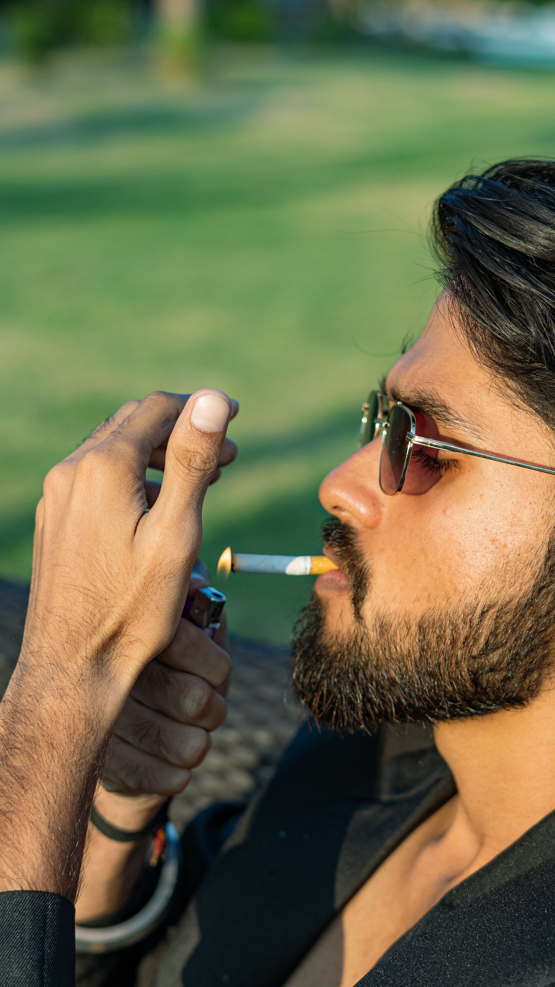 Man wearing sunglasses lighting a cigarette outdoors on a sunny day.