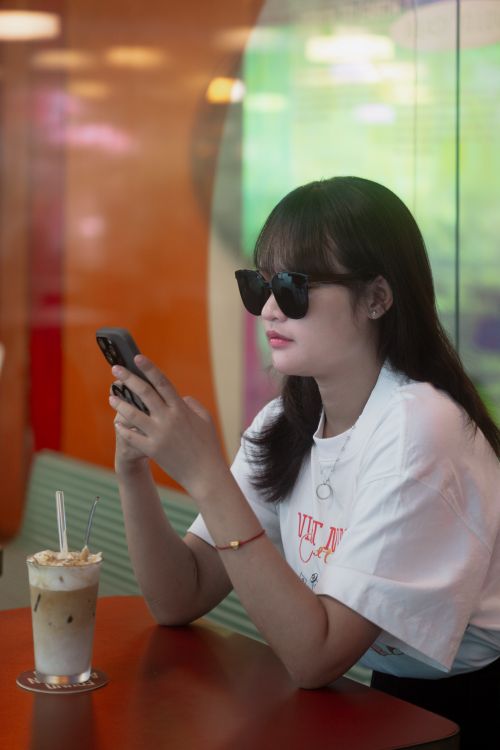 Free Young woman in sunglasses enjoying a drink while using smartphone in a modern cafe setting. Stock Photo