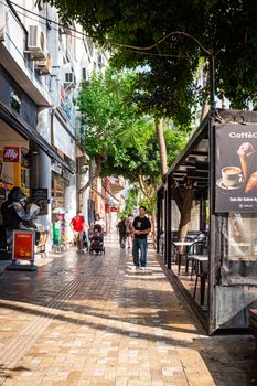Bustling city street with people walking, shops, and outdoor seating under bright sunlight.