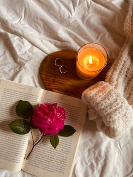 Warm cozy setup with candle, book, and pink rose on a bed.