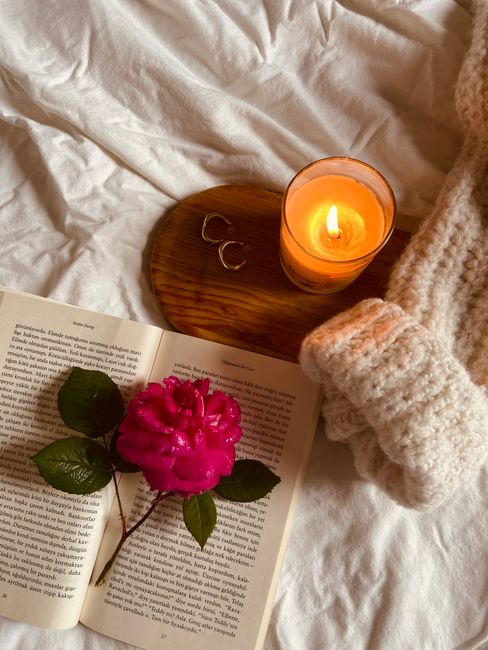 Warm cozy setup with candle, book, and pink rose on a bed.