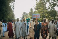 A vibrant cultural procession with a diverse group holding a 'Dan Majen' sign outdoors.