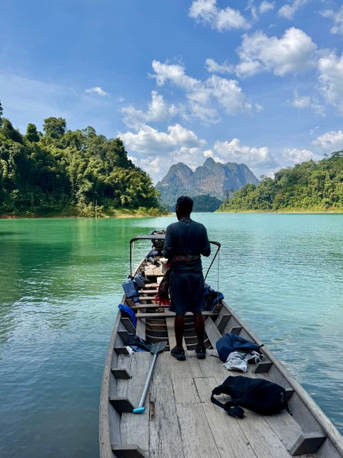 Free Man steering boat on tranquil emerald lake surrounded by lush mountains under clear blue sky. Stock Photo