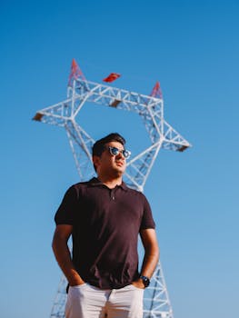 Man in sunglasses stands confidently before a geometric power tower on a clear day.