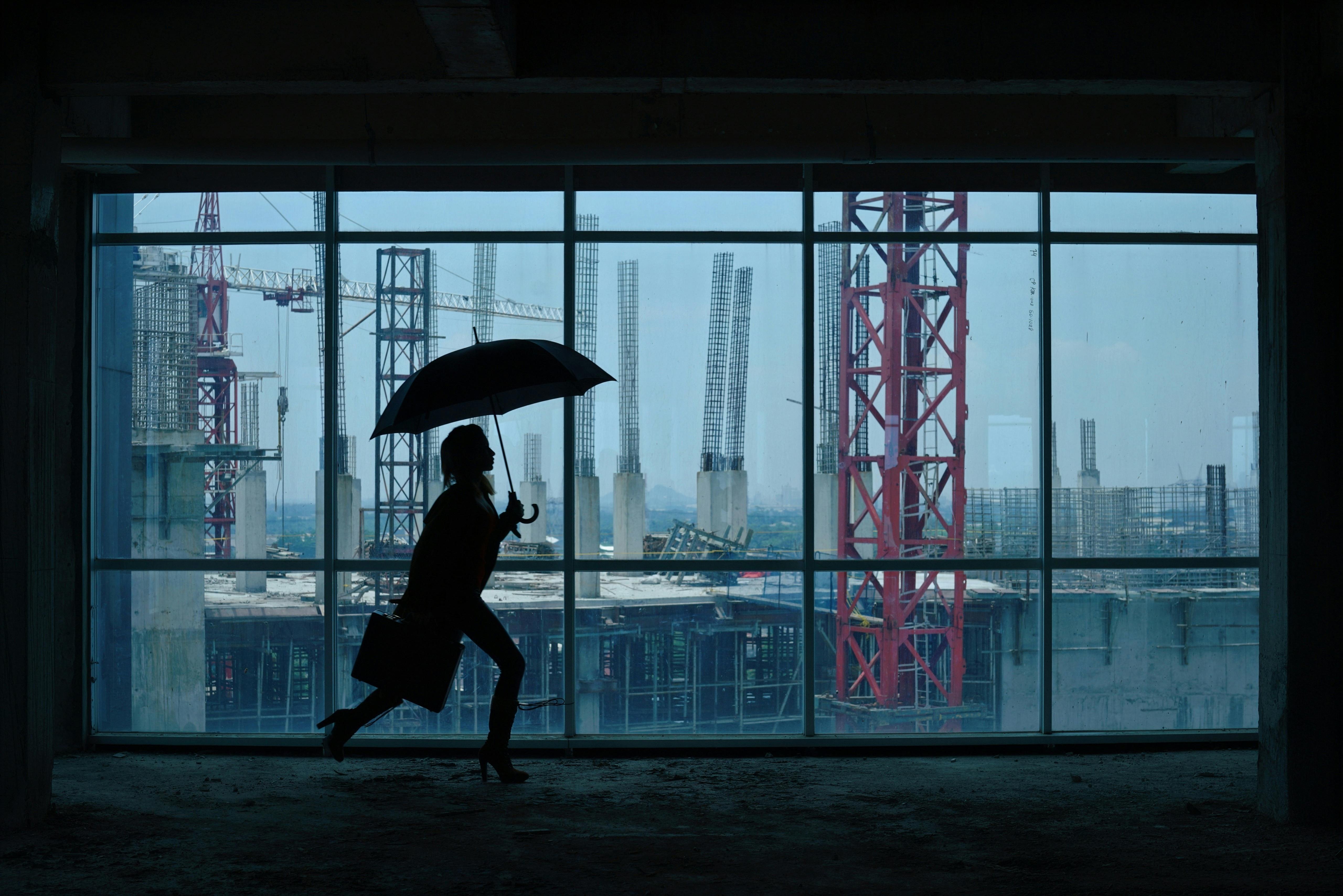 Silhouette of person with umbrella and briefcase running inside a building, overlooking construction site through large windows.