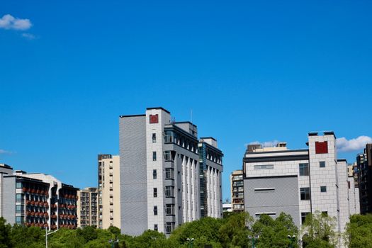 A vibrant cityscape featuring modern buildings under a bright blue sky, lush greenery at the base.