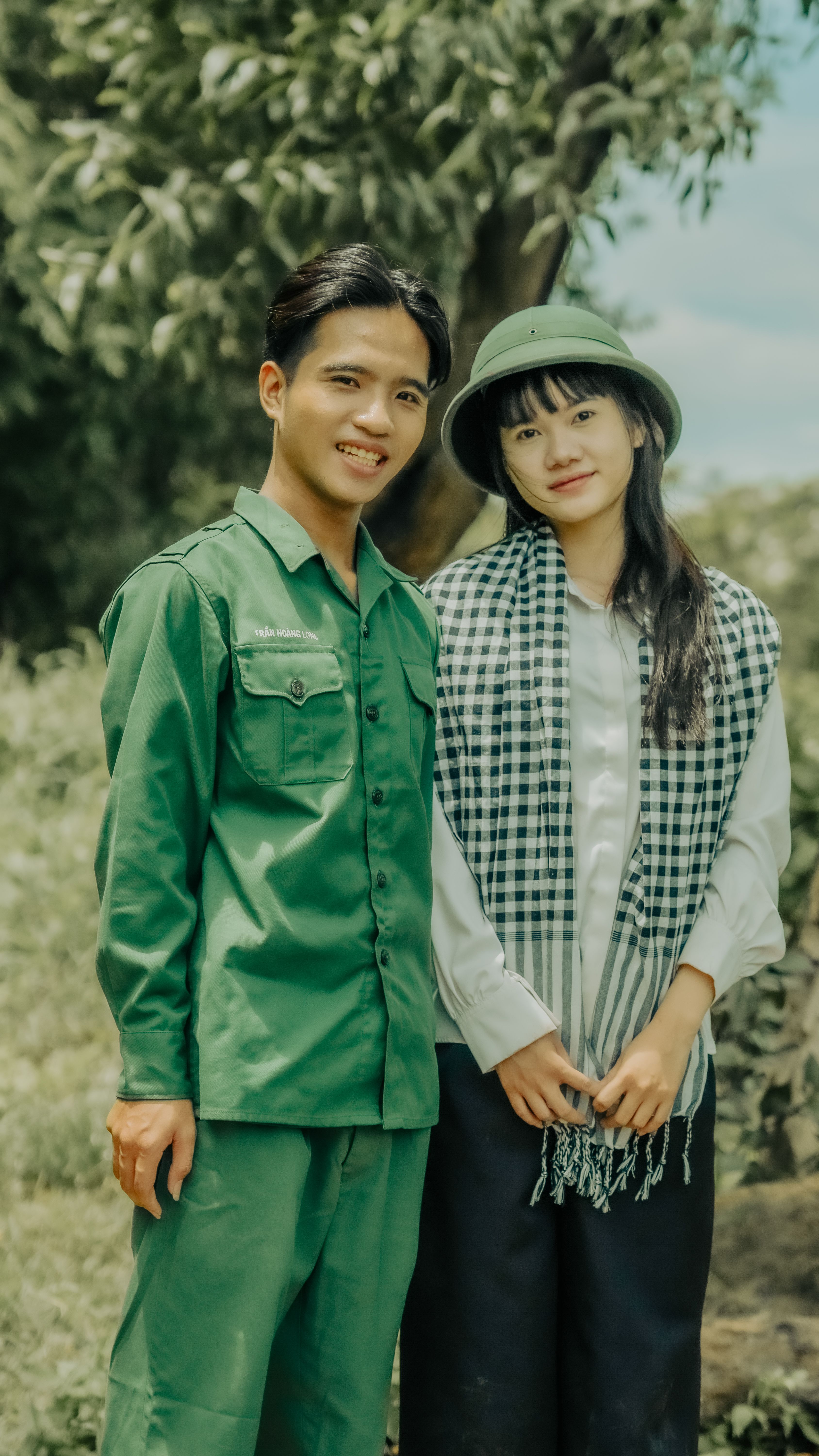 Smiling young couple in traditional attire posing outdoors with lush greenery.