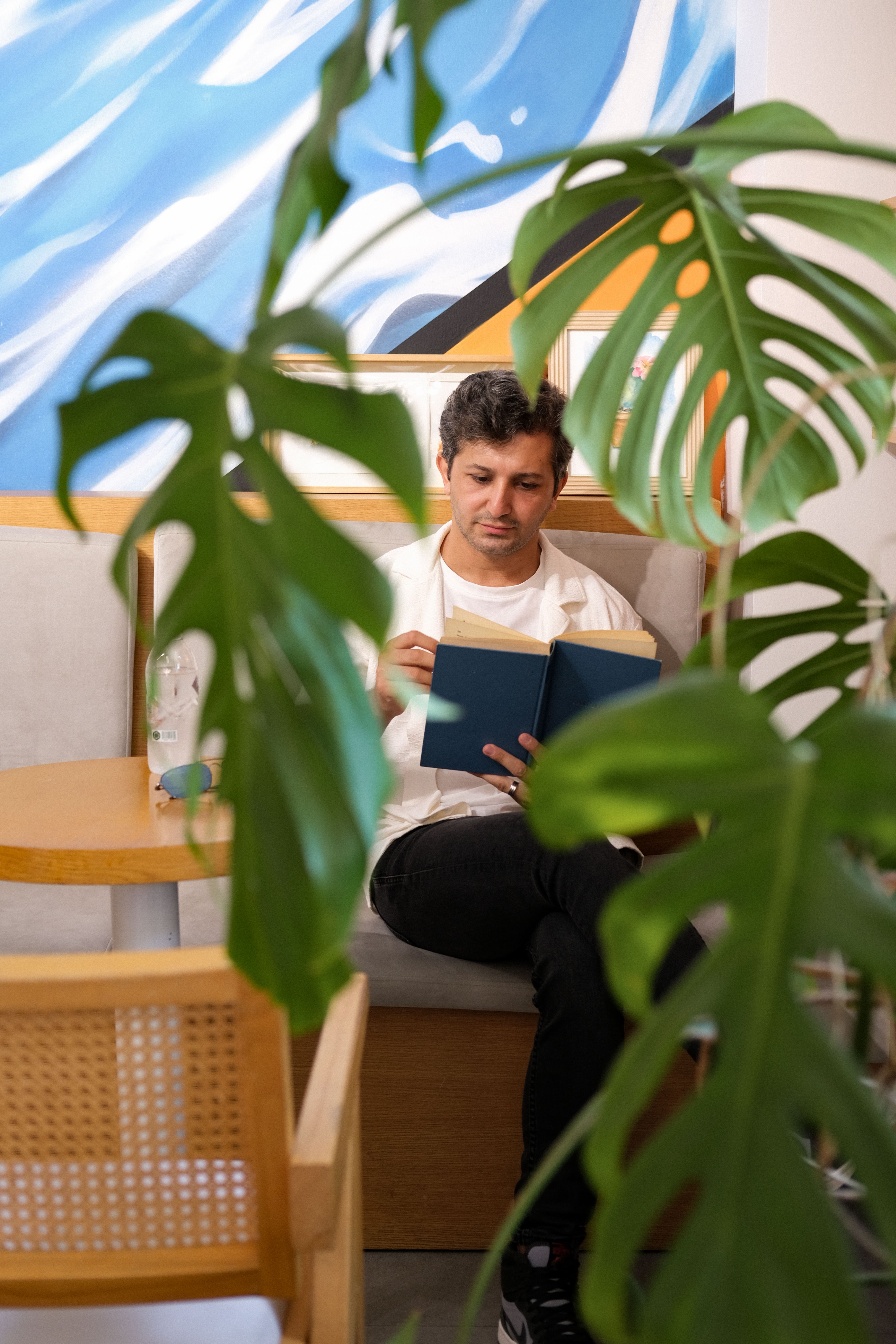 A man enjoys reading in a cozy corner of a café, surrounded by plants.