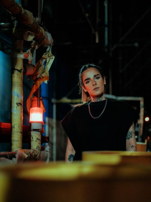 Free A young woman with tattoos stands confidently in an industrial environment at night, surrounded by pipes and a red warning light. Stock Photo