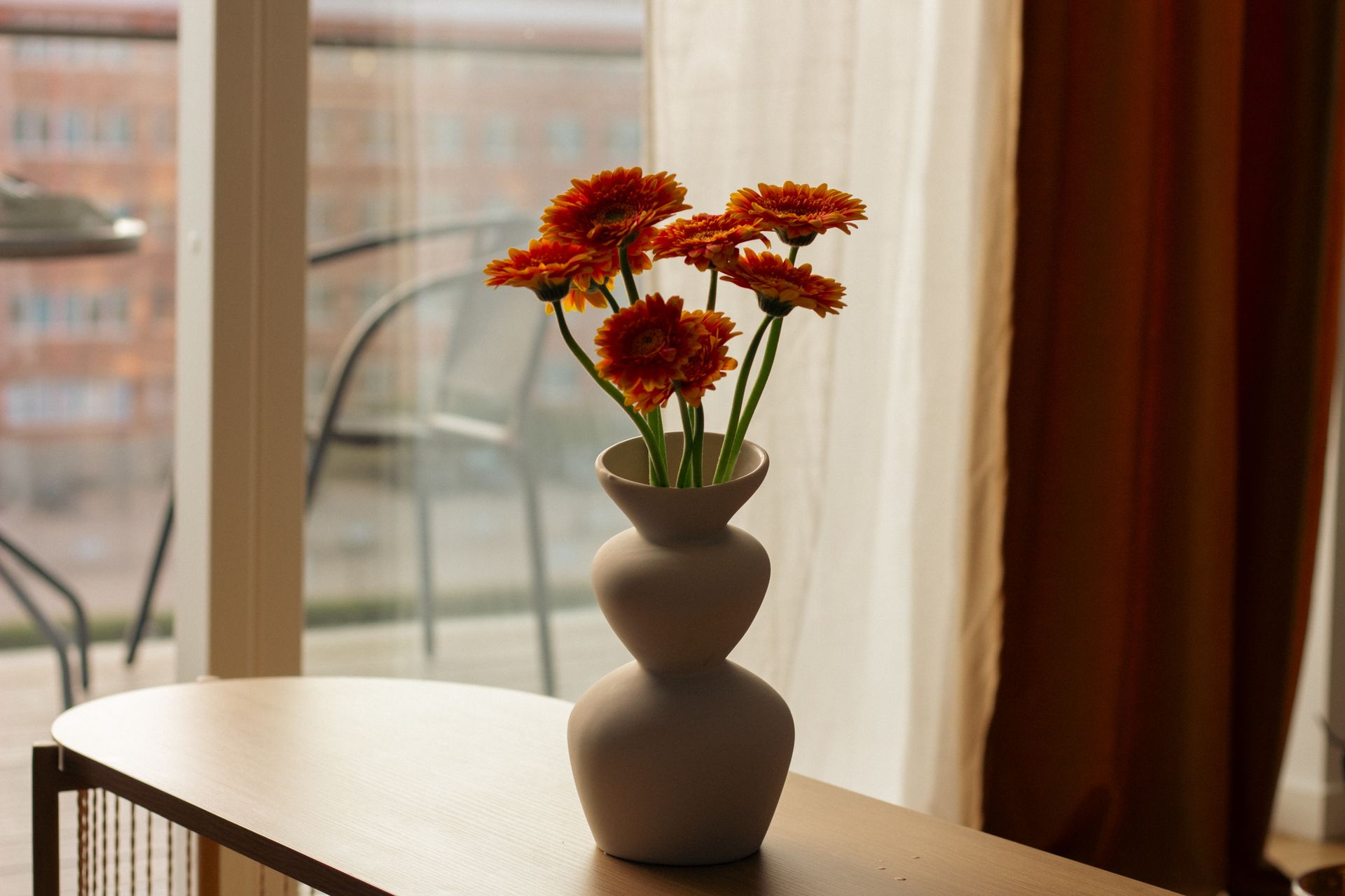 A modern vase holds bright orange flowers on a sunlit indoor table.