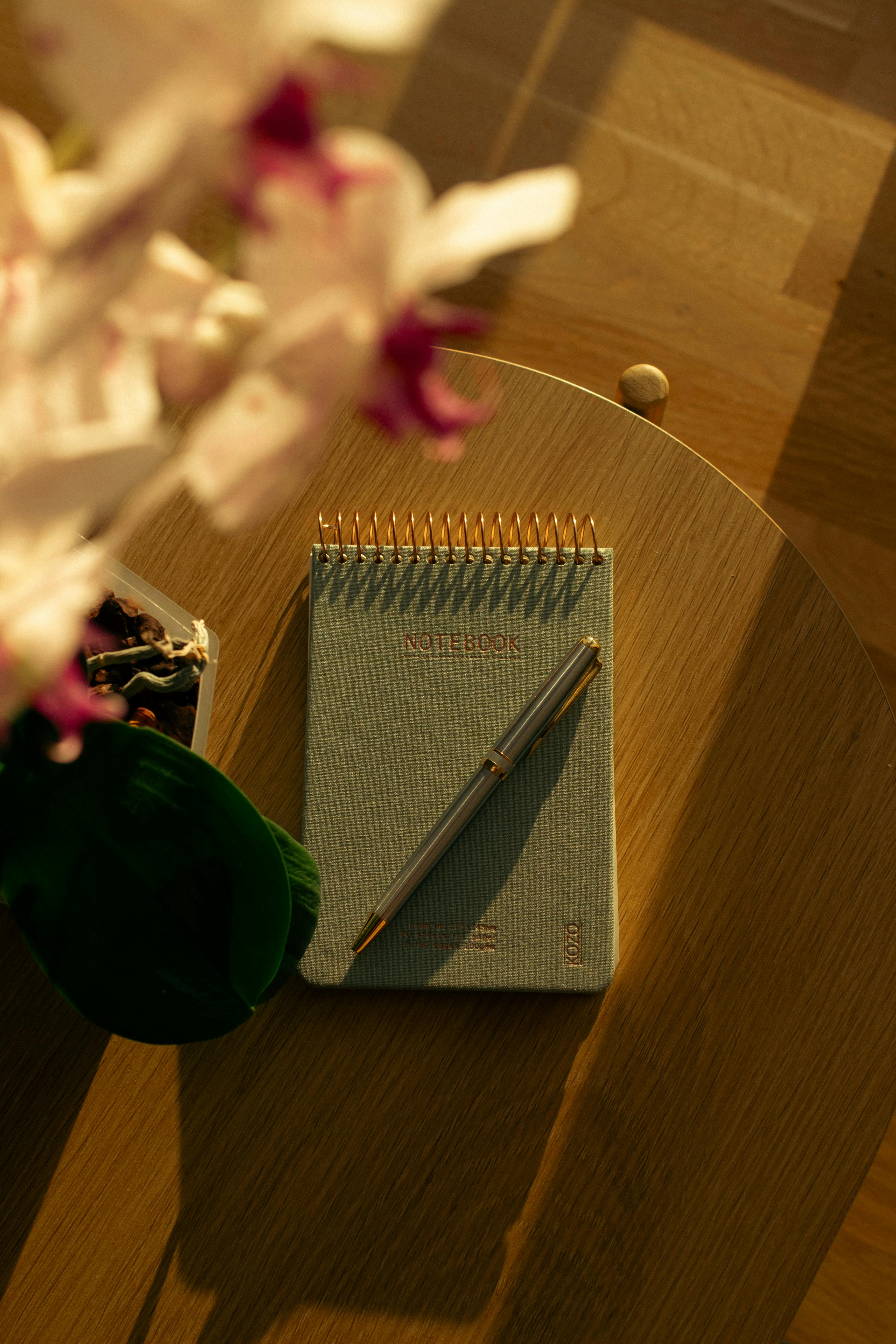 Gratuit Blocnotes spiralat însorit cu pix pe o masă de lemn lângă o orhidee înflorită. Fotografie de stoc