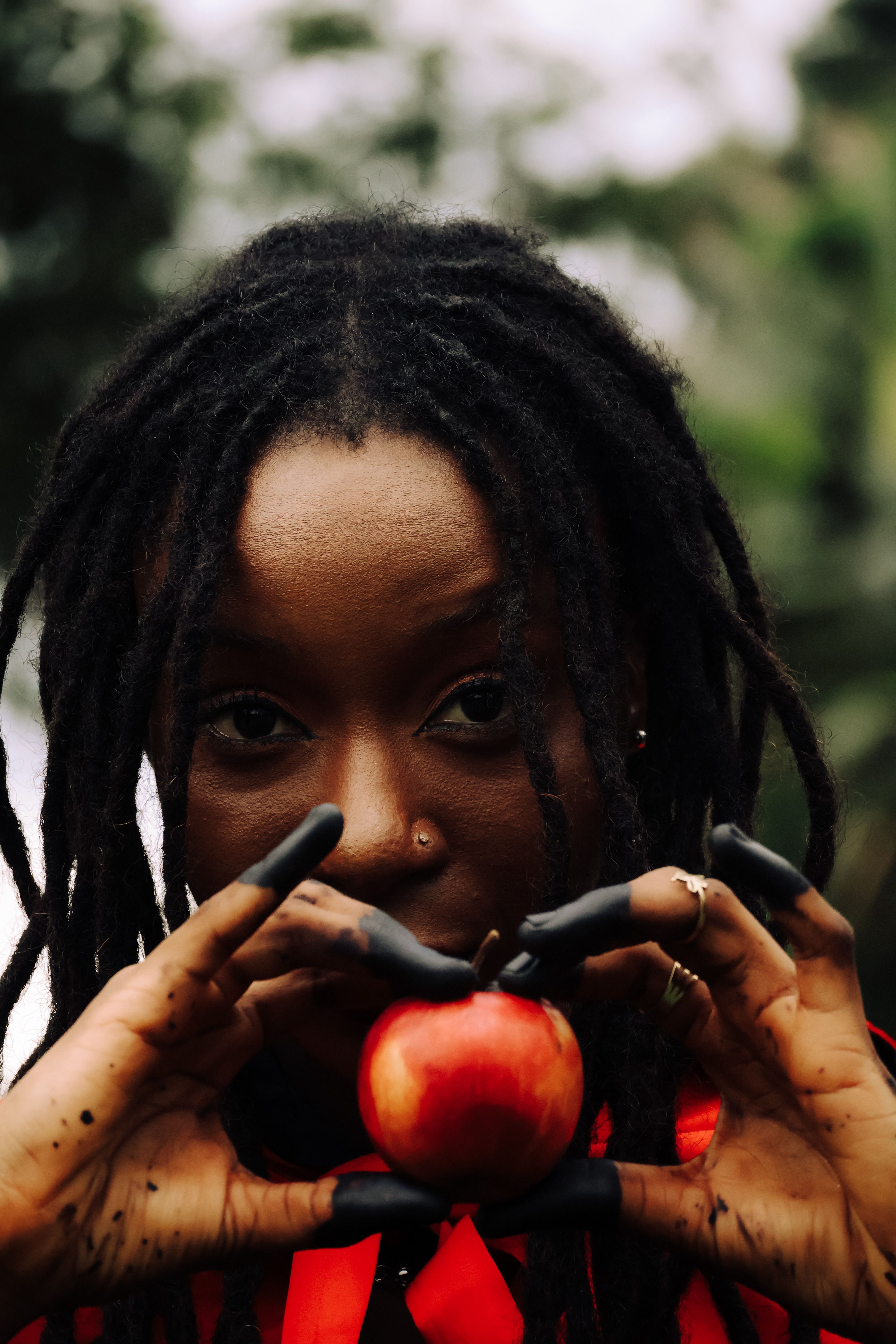 A woman with dreadlocks holds a red apple outdoors, evoking natural themes.