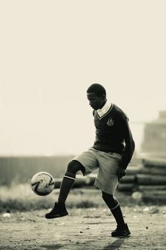 A teenage boy skillfully juggling a football on a dusty field in Kampala, Uganda.