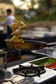 A chef skillfully tosses potato wedges in a frying pan over a stove in an outdoor kitchen setup.