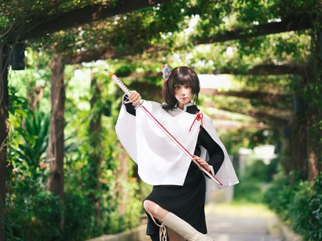 A cosplayer in an anime-inspired outfit poses outdoors with a sword, surrounded by greenery.
