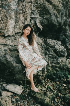 A woman in a floral dress poses gracefully against a rugged rock. Outdoors beauty.