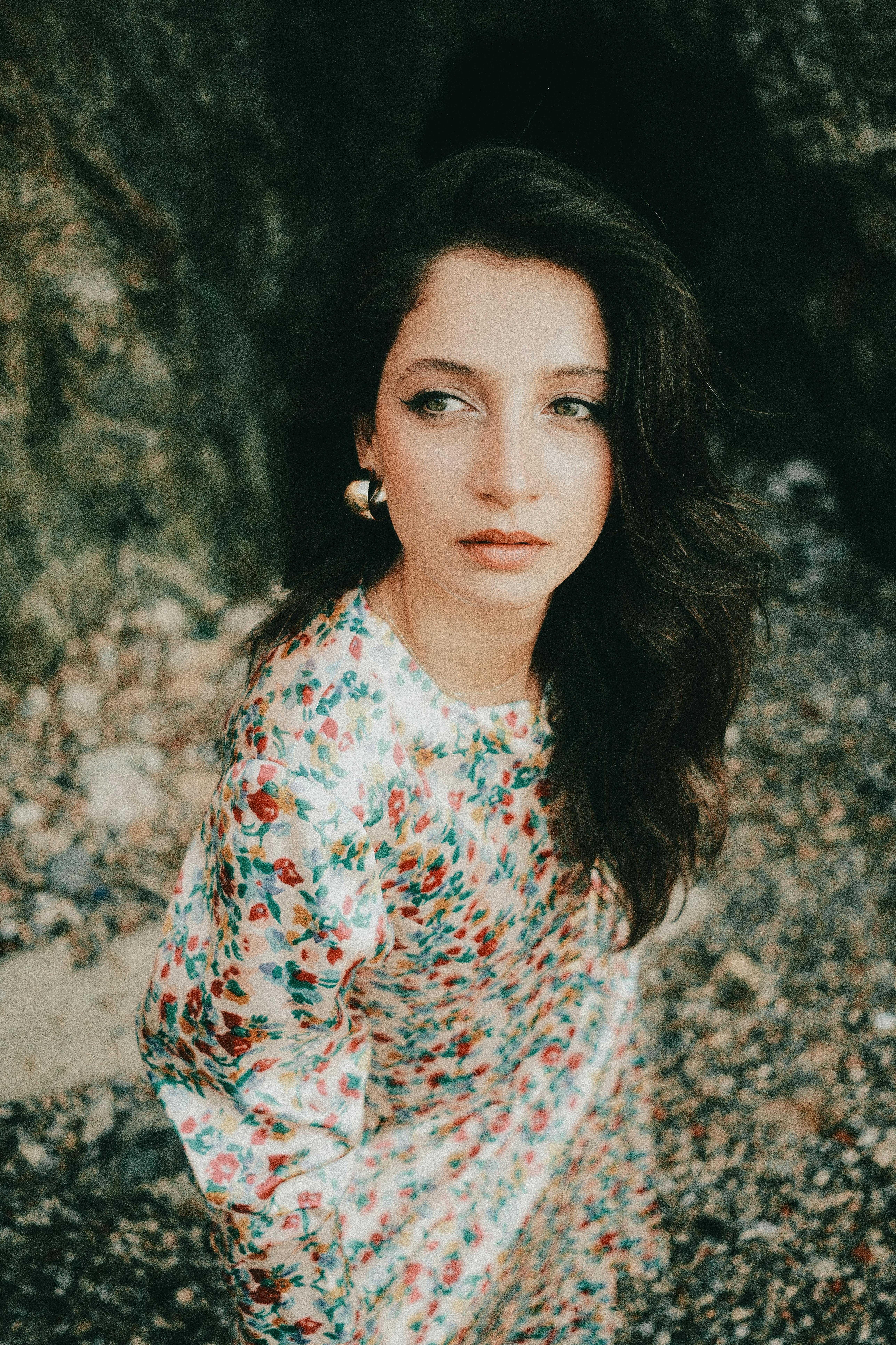 Elegant woman in floral dress standing by rocky terrain, exuding a serene and introspective mood.