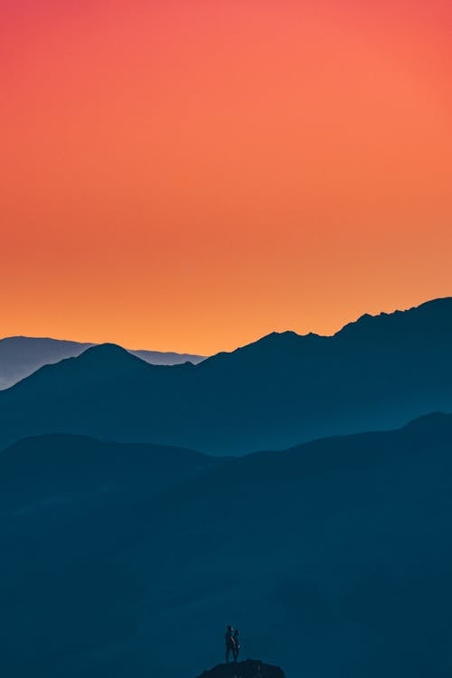 Free Silhouette of unrecognizable hikers standing on top of rocky cliff surrounded by high mountains against amazing colorful sunset sky Stock Photo
