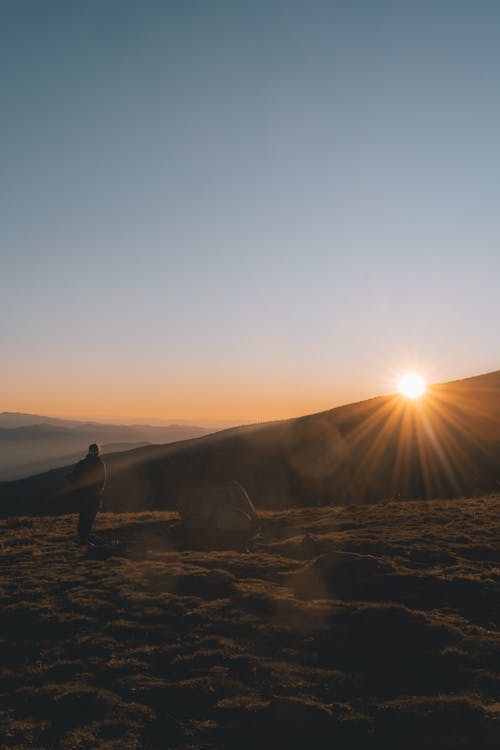 Anonymous traveler standing on rough rocky terrain near mountain range against cloudless blue sky during sunset