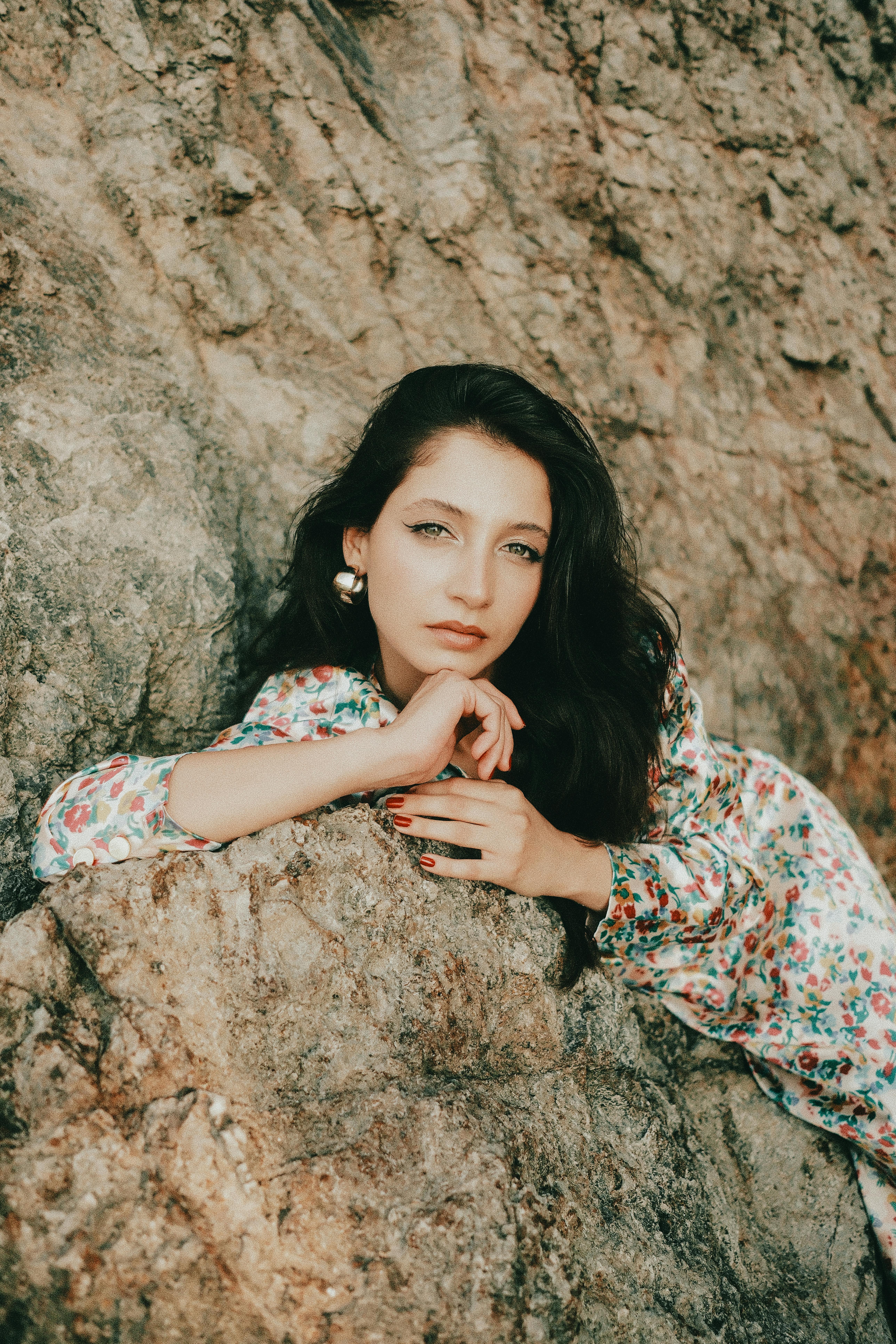 Stylish woman in floral dress posing against a rocky surface outdoors.