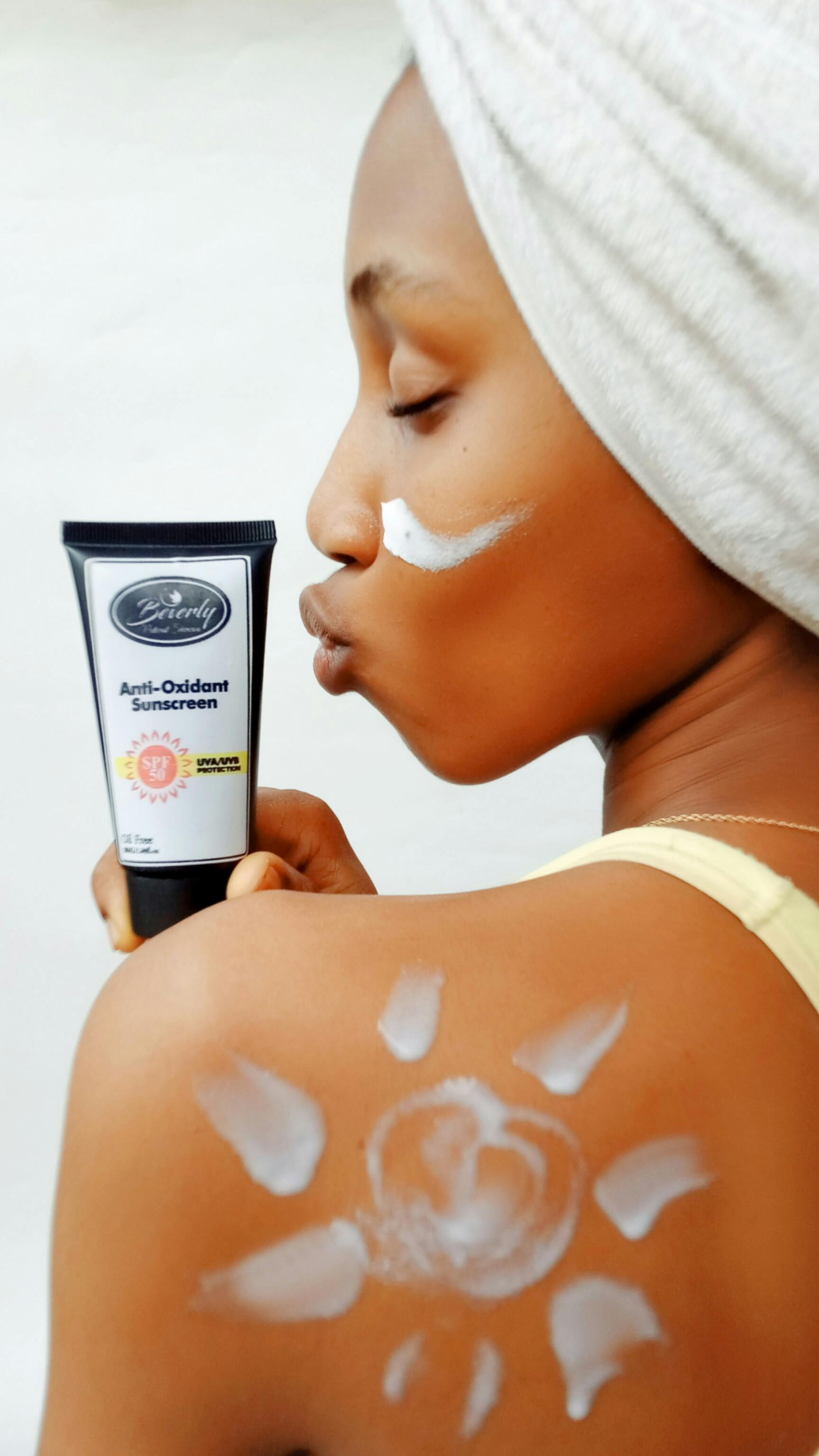 Woman with towel on head applying sunscreen on shoulder, holding tube.