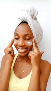 A woman with a towel on her head applies facial cream, showcasing a skincare routine.