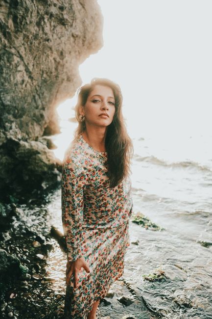 A young woman in a floral dress standing by the rocky seaside during sunset.