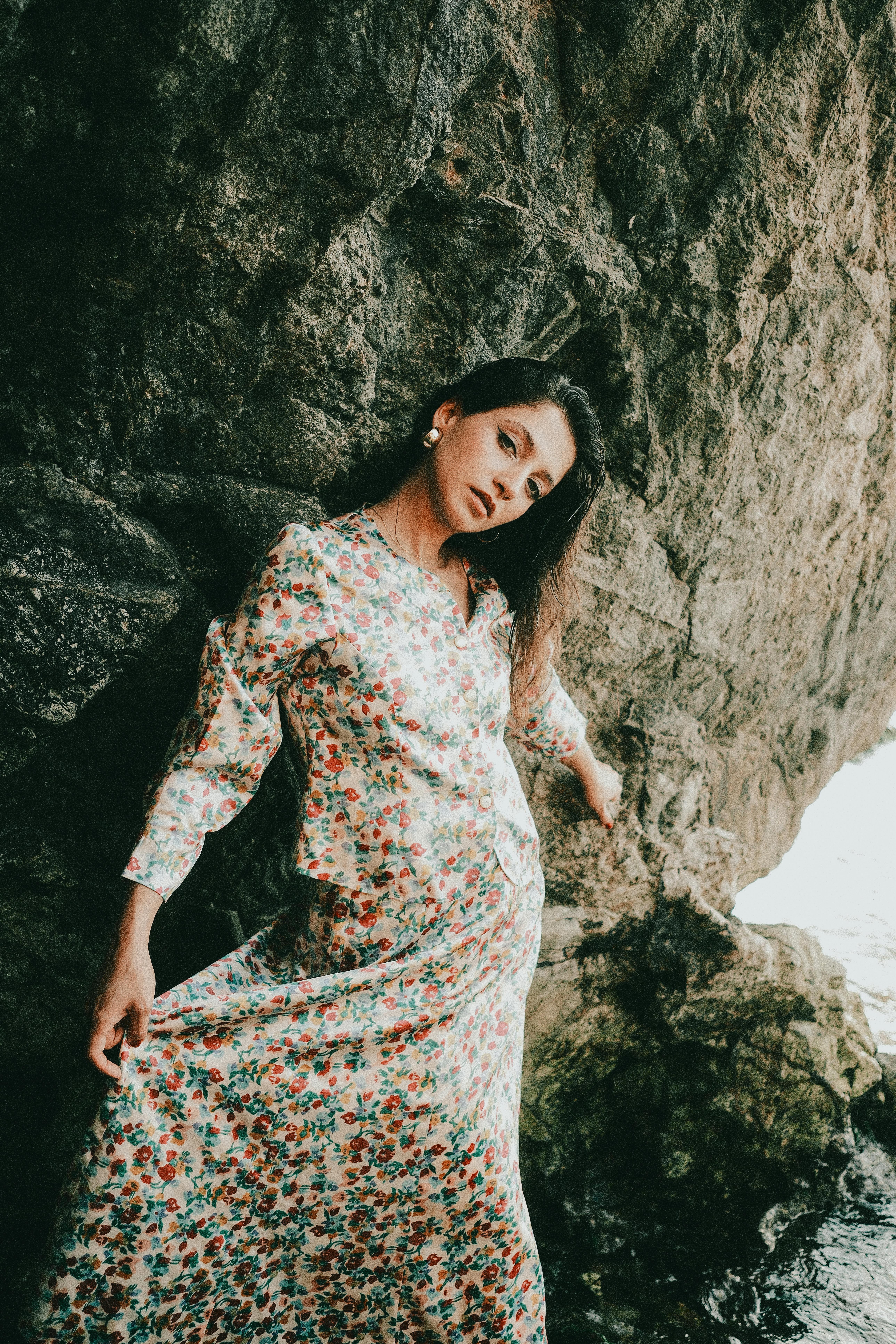 Elegant woman in a floral dress leaning against a rocky cliff outdoors.