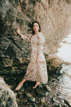 A woman in a floral dress stands beside rocky seaside, exuding grace and poise.