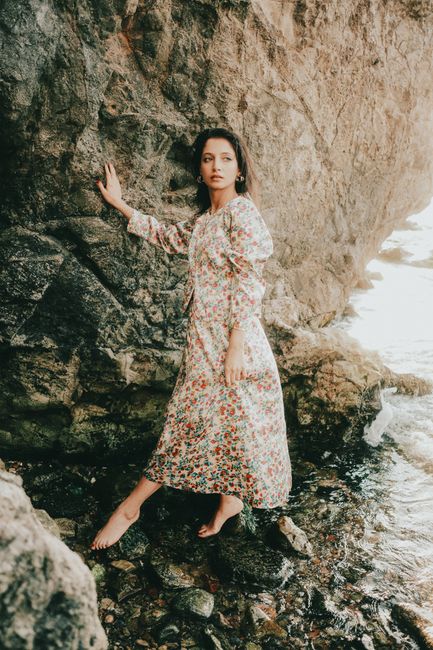 A woman in a floral dress stands beside rocky seaside, exuding grace and poise.