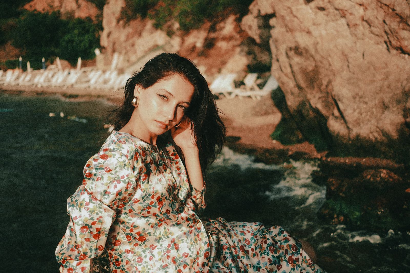 Woman in a floral dress sitting on rocks by the sea in warm golden sunlight
