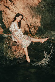 Smiling woman in floral dress sitting on rocks by the sea, enjoying summer.