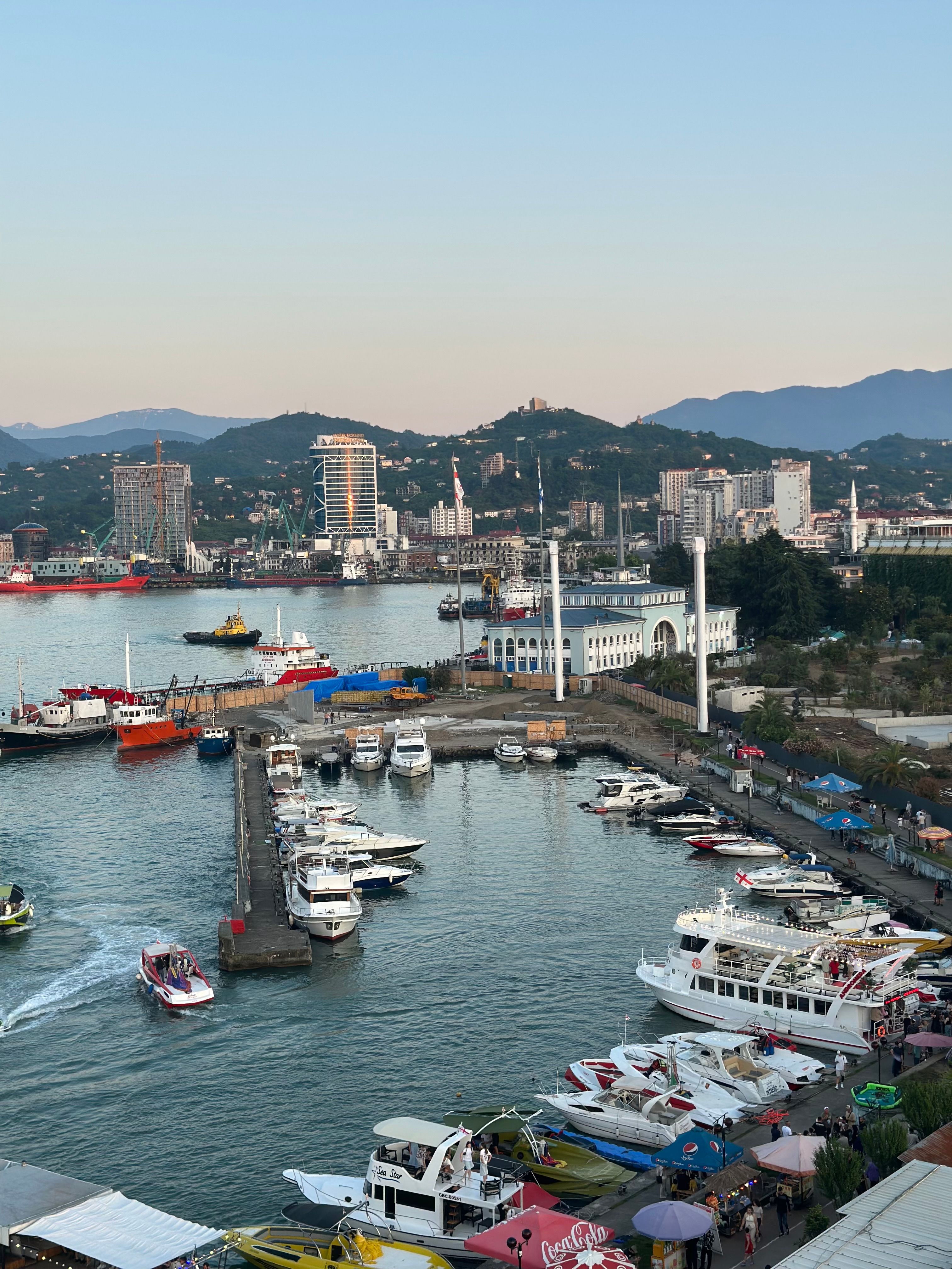 A bustling harbor with boats against a cityscape at sunset, surrounded by hills.