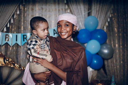 A joyful woman holds her child at a festive indoor birthday party with balloons.