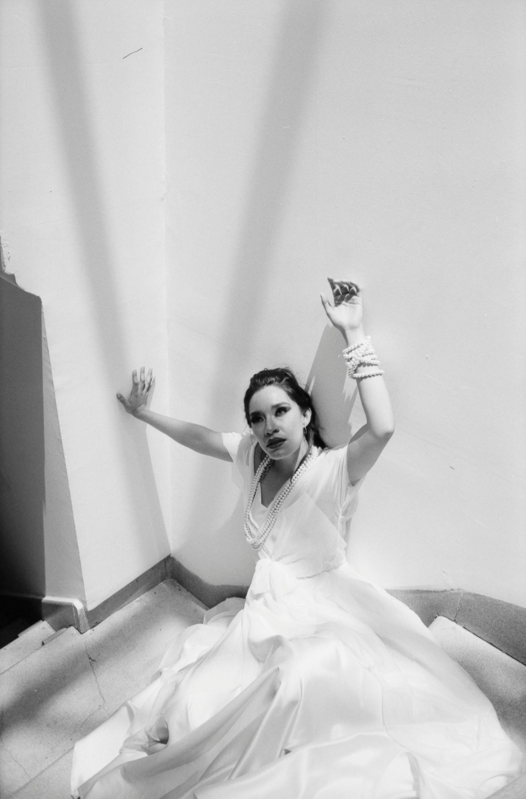 Gratis Una donna in abito bianco posa in modo teatrale contro un muro bianco, trasudando eleganza ed emozione. Foto a disposizione