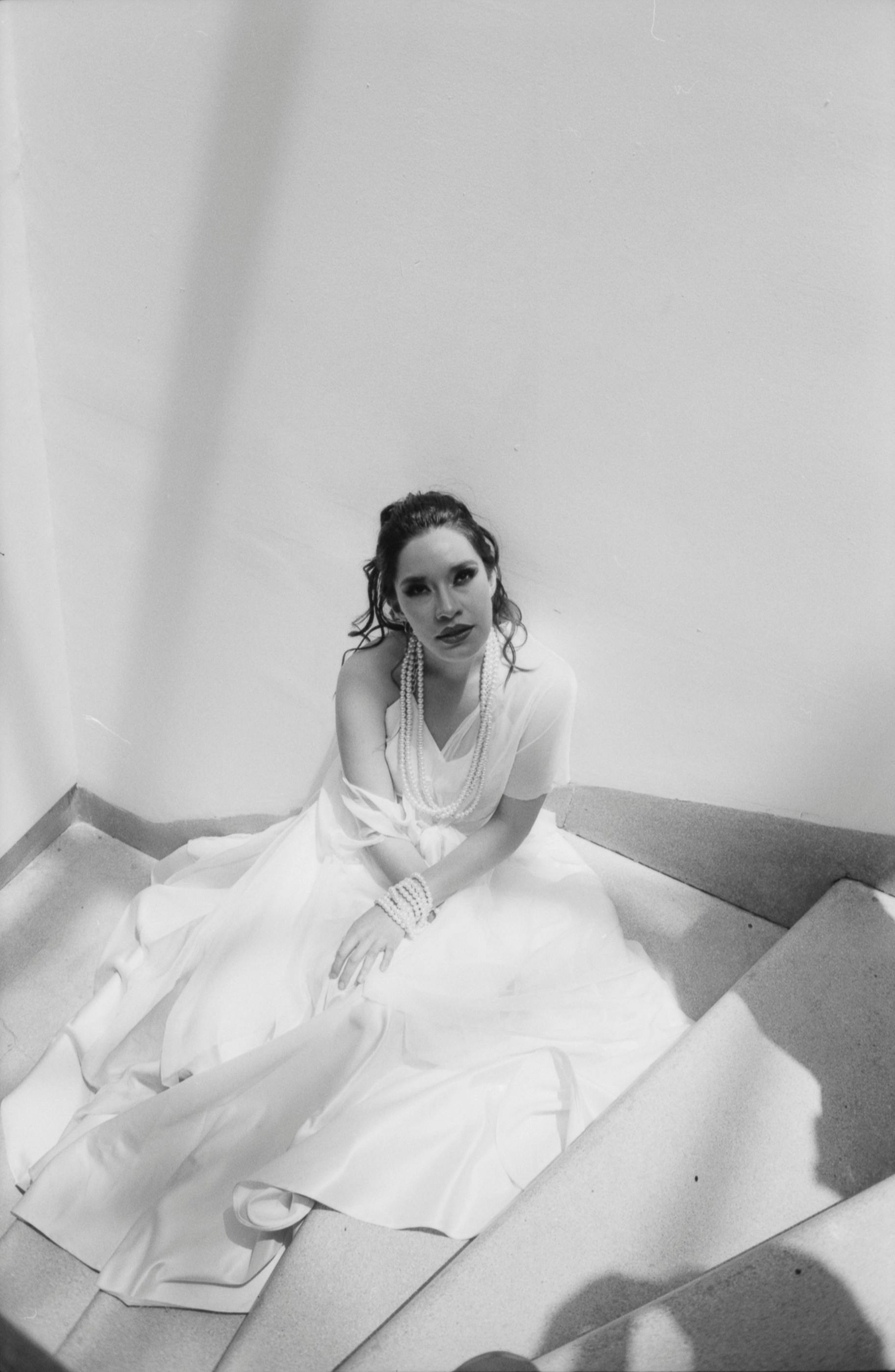 Gratis Una donna elegante, con un abito bianco svolazzante, è seduta elegantemente sulle scale, immersa in una luce soffusa. Foto a disposizione