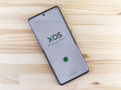 Modern smartphone showing XOS interface placed on a light wooden surface, showcasing technology and design.