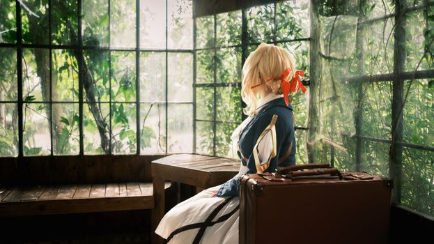 Cosplay character in vintage outfit with suitcase in rustic wooden room with large windows.