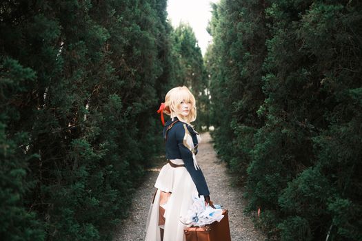 A cosplayer in fantasy attire walks through a verdant forest path, holding a brown suitcase.