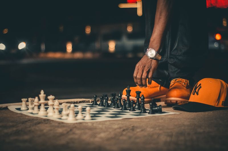 A person sets up a chess game on a city street at night, featuring stylish footwear and vibrant cap.