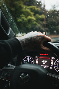 Close-up of a tattooed hand on a steering wheel inside a car with rain outside the window.