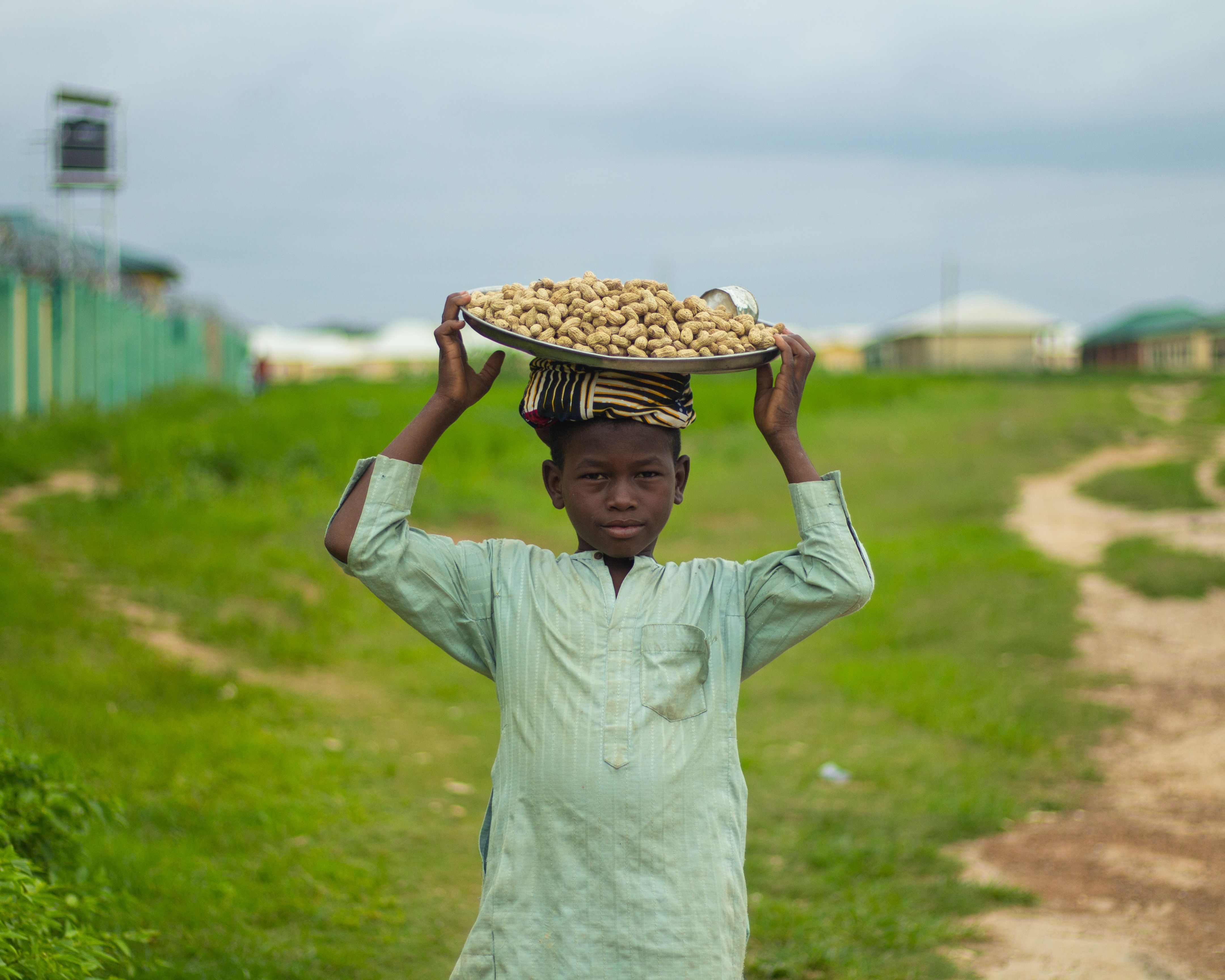 Young boy in traditional attire carries tray of peanuts outdoors.
