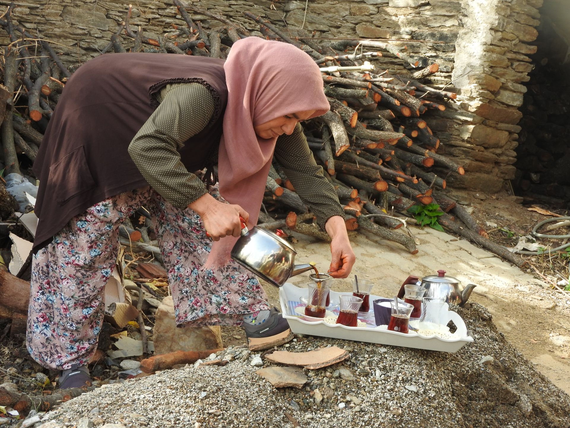 An elderly woman in a headscarf pours tea outdoors, showcasing cultural tradition using glassware and a metal kettle.