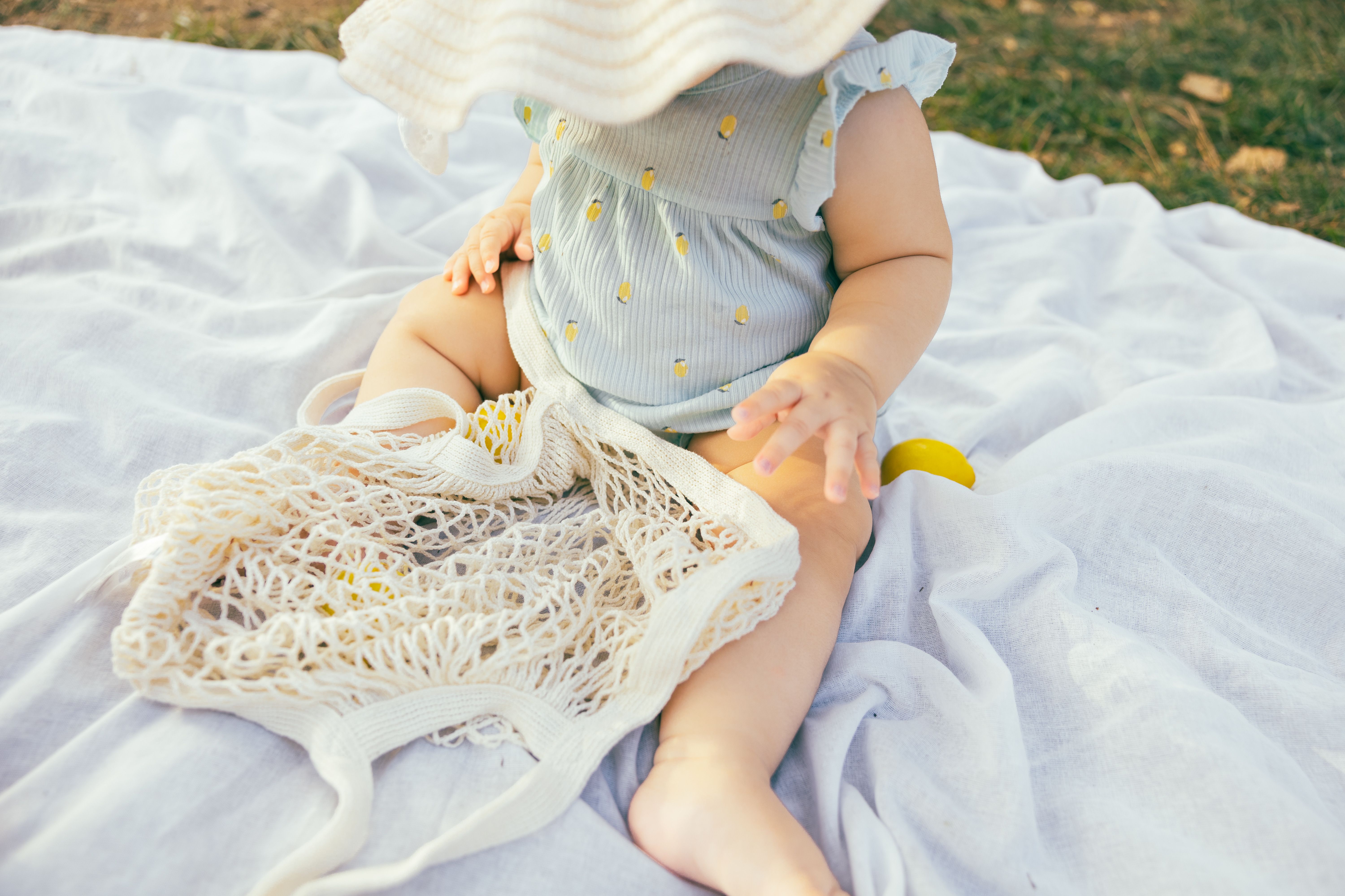 Adorable baby in summer outfit with sun hat and net bag on a picnic blanket.