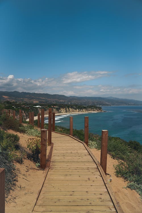 From above wooden walkway on hilly terrain leading to sandy ocean beach against cloudy blue sky