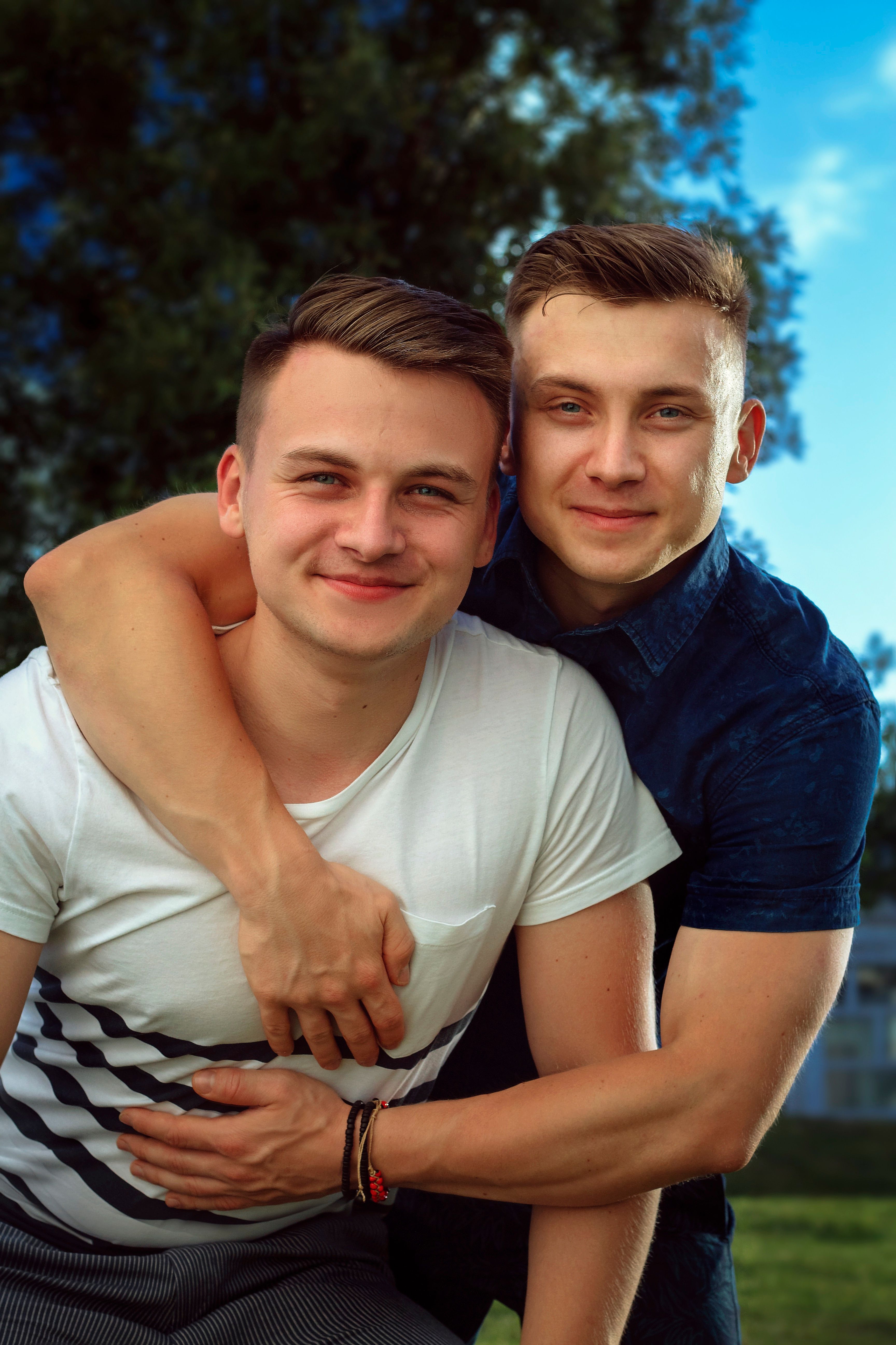 Two young men smiling warmly, embracing outdoors on a sunny day.