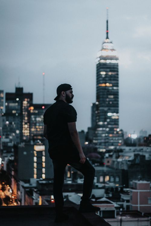 Free Man standing on rooftop gazing at illuminated city skyline during twilight, urban exploration. Stock Photo