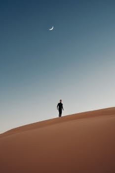 A lone person walks across desert dunes under a crescent moon at Siwa Oasis, Egypt.