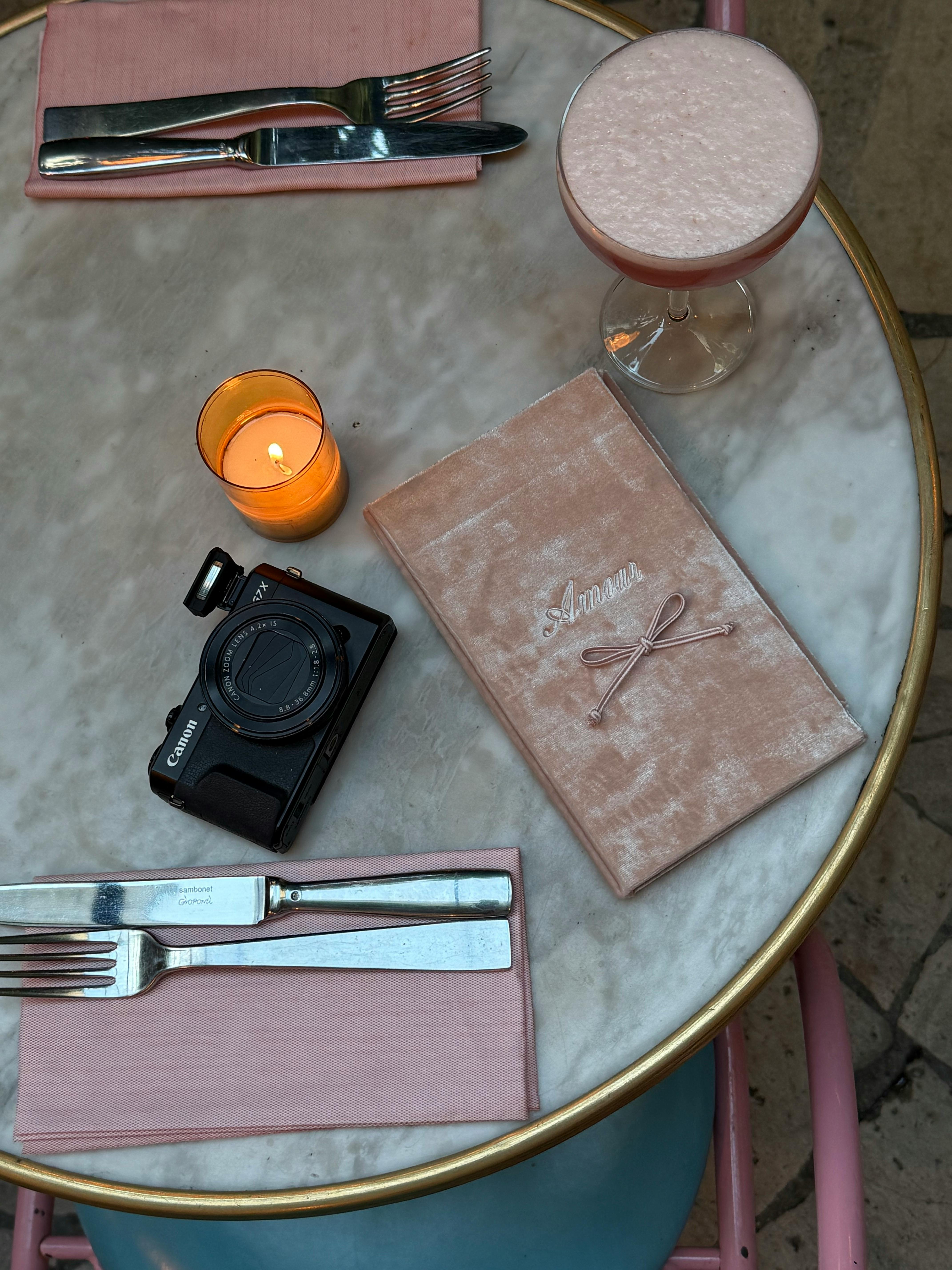 Stylish table setting featuring a menu, camera, candle, and drink on a marble surface.